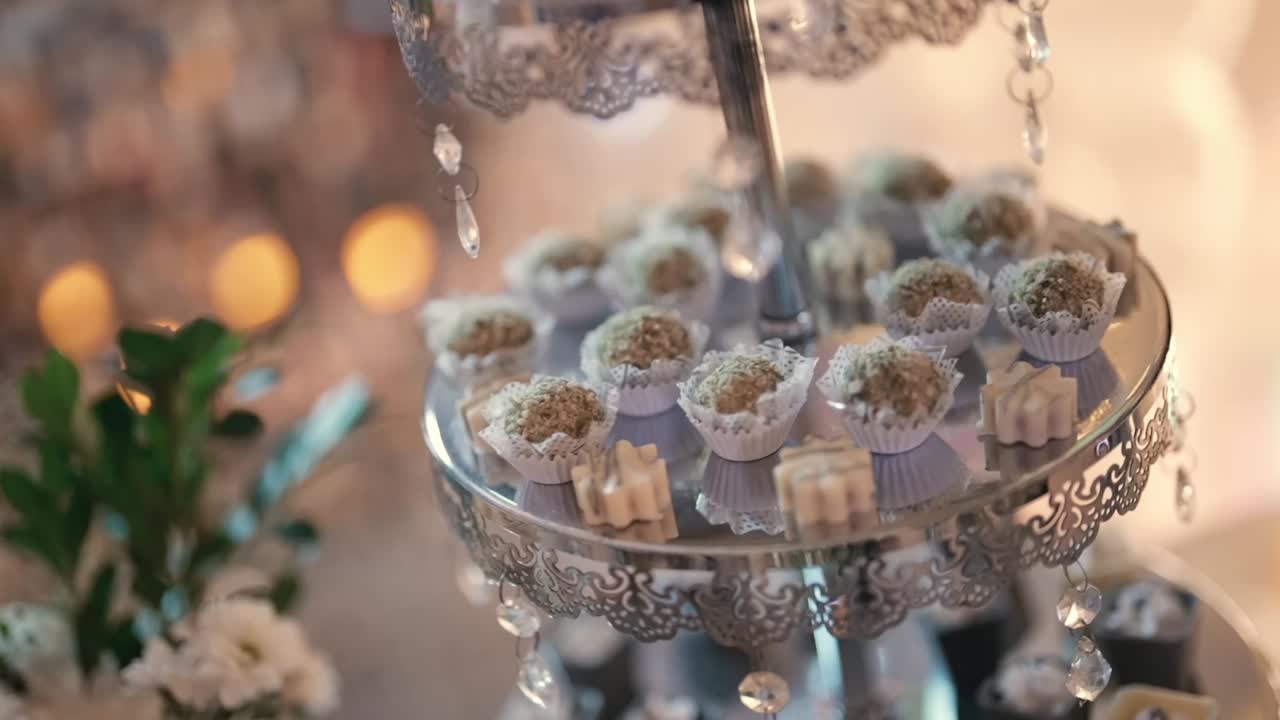 Slow motion-afsløring af et elegant dessertbord ved et luksusarrangement viser et udvalg af appetitvækkende desserter i bidder arrangeret på et krystalstativ med varmt bokeh-lys, raffineret dekoration og lav dybdeskarphed.