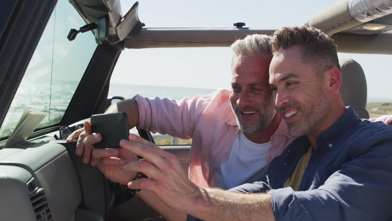 feliz pareja de hombres gays caucásicos tomando selfies sentados en el coche sonriendo en un día soleado en la playa