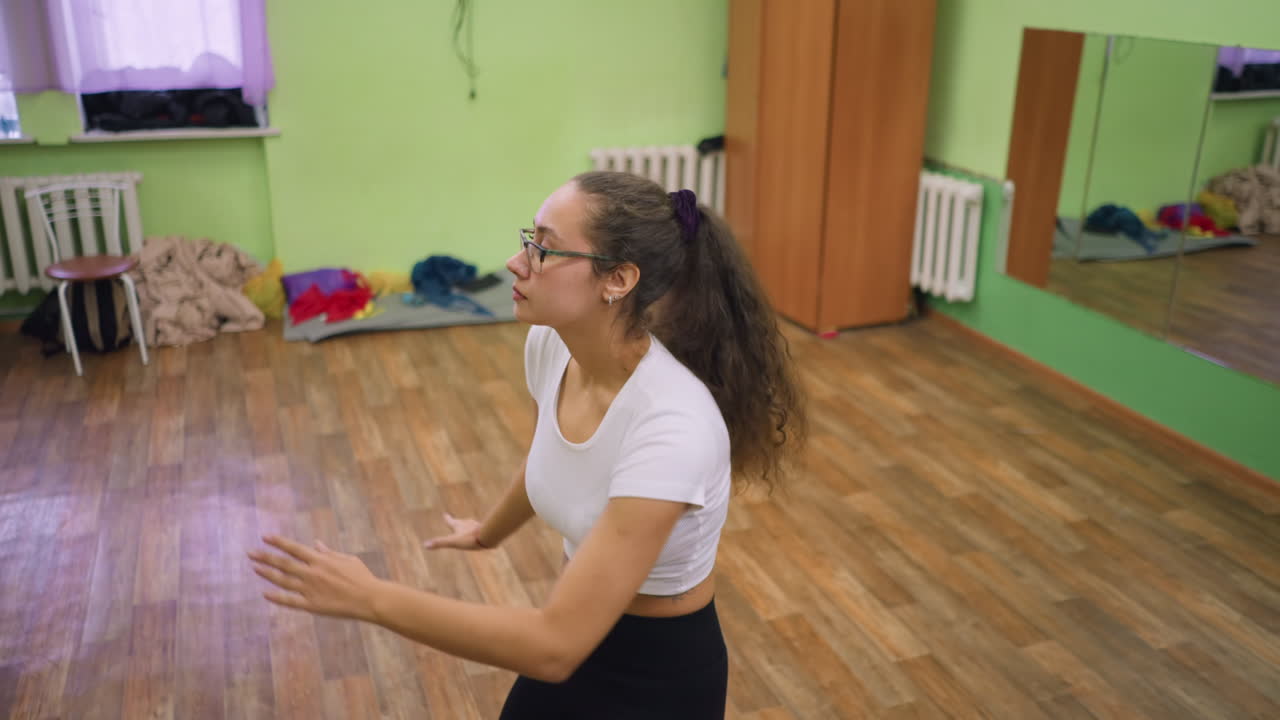 Pretty lady in white top dancing in room with mirror reflecting her back, red lace visible on wrist, wooden floor adding warmth to setting, movement showing energy, focus and expressive confidence