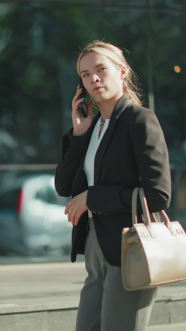 Side view of banker on phone with customer showing frustration during tense conversation while walking past mall with glass facade reflecting cars, trees, and passerby entering entrance