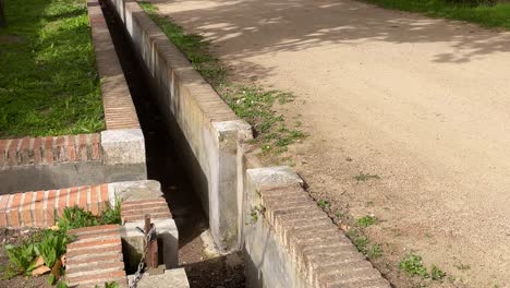 there is an irrigation canal made of brick and mortar on the side of a dirt road on one side there are cypresses and on the other side there are various trees jardines de el principe aranjuez spain