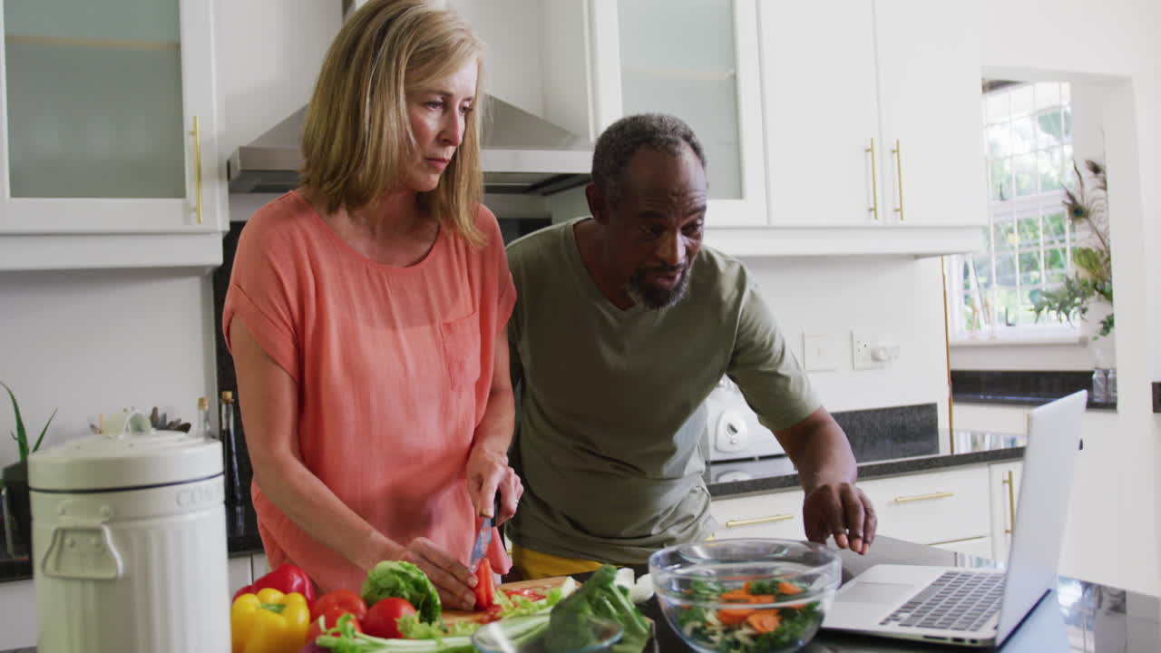 Diverse senior couple preparing food in kitchen following recipe on laptop