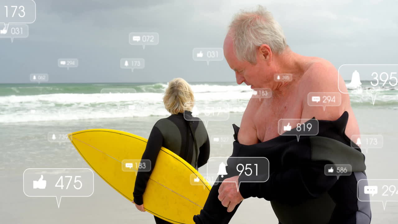 Senior couple pulling wetsuit and holding surfboard at beach with floating social media icons