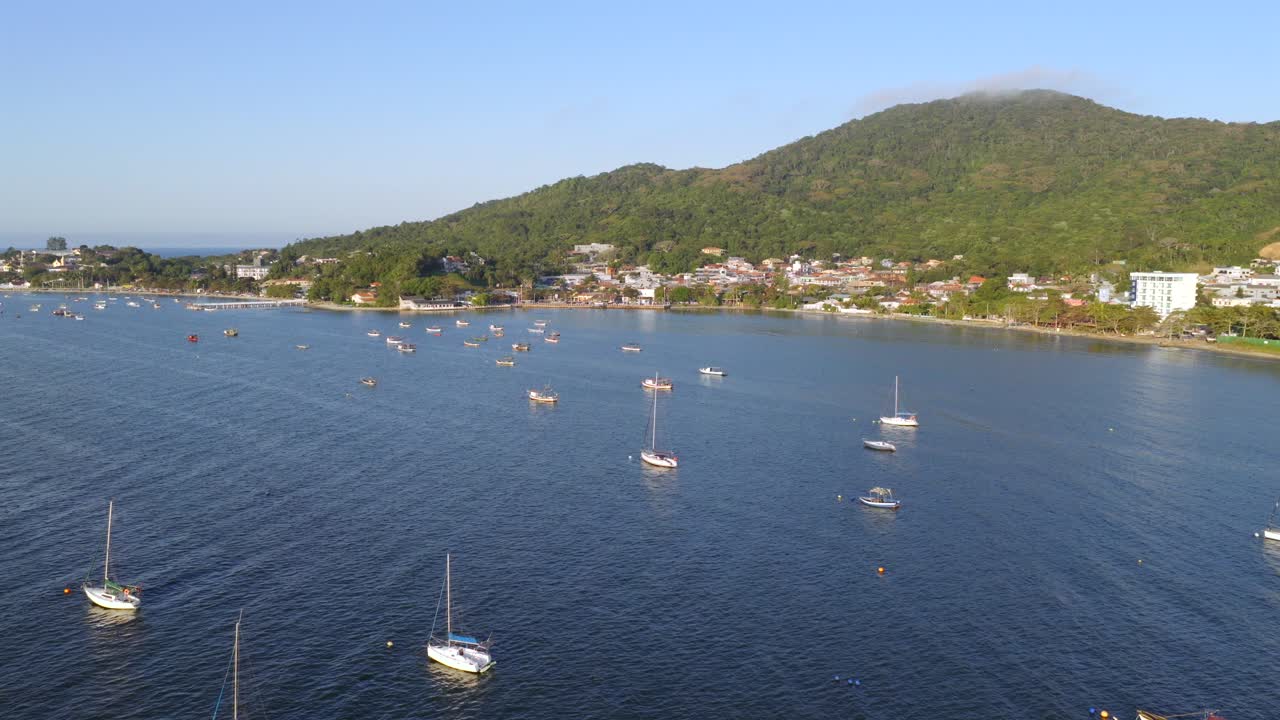 Drone clip of moored sailboats seen from offshore, with coastal homes and green hills behind. Forward shot in afternoon