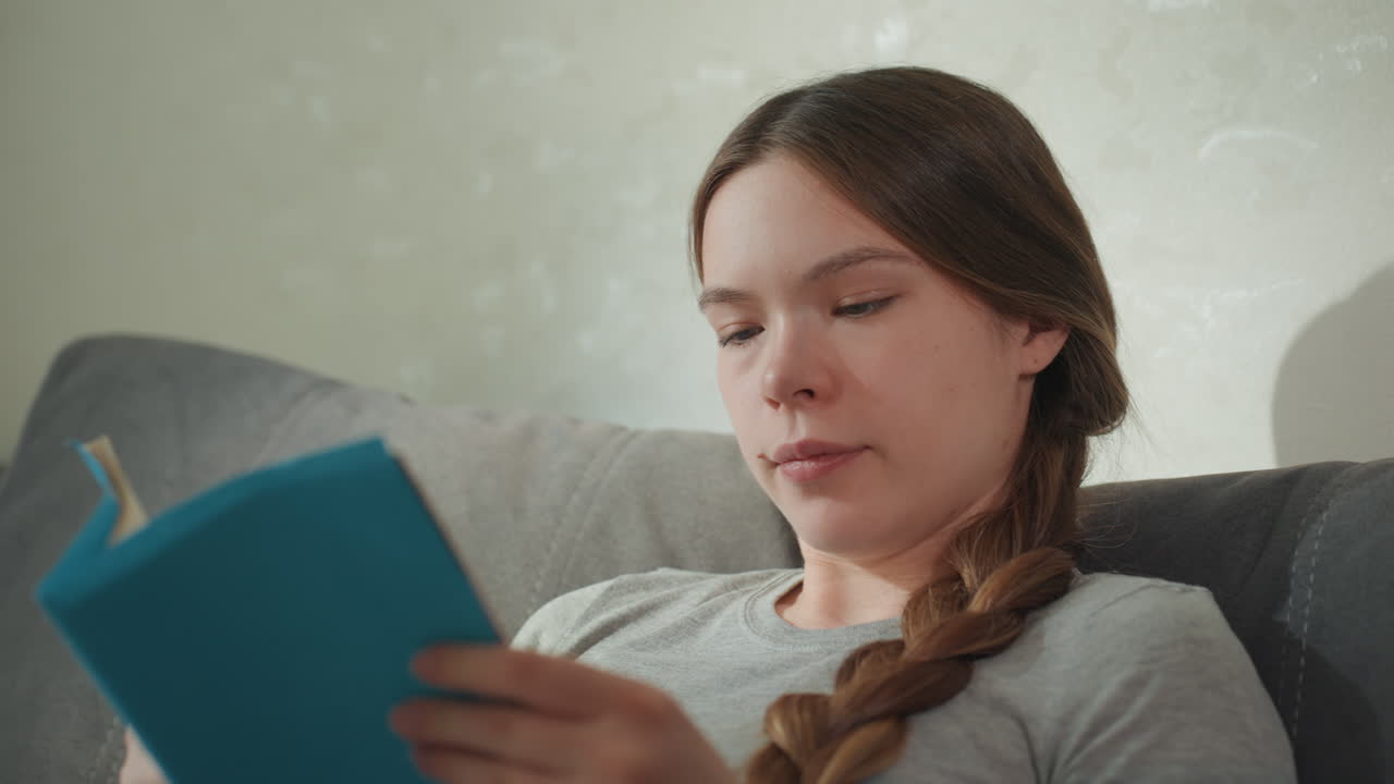 Woman reading blue book on couch with attentive expression, closeup sequences show concentration and page turning, braided hair and soft daylight create studious home atmosphere, portraying study