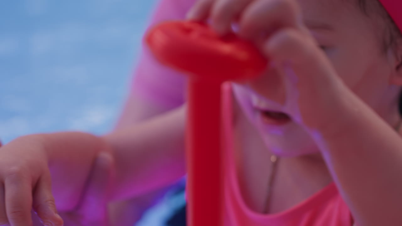 Close view of baby placing red ring toy onto foam stick, tiny hands stacking play near pool, calm learning moment with bright color contrast, early coordination building, gentle support off camera