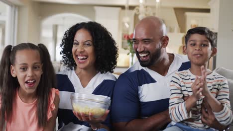 Happy-biracial-parents,-son-and-daughter-watching-sport-on-tv-with-popcorn-at-home,-slow-motion