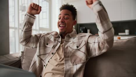 a happy and very cheerful young man with black skin and brunette is very happy about his success at work sitting at a laptop on the sofa his girlfriend runs up to him and hugs him from behind rejoicing for her boyfriend in a modern apartment