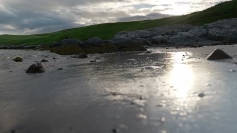 A-low-angle-shot-of-waves-gently-lapping-up-against-a-sandy-beach-very-close-to-the-camera-as-the-sun-sets-behind-grassy-hills-in-the-background