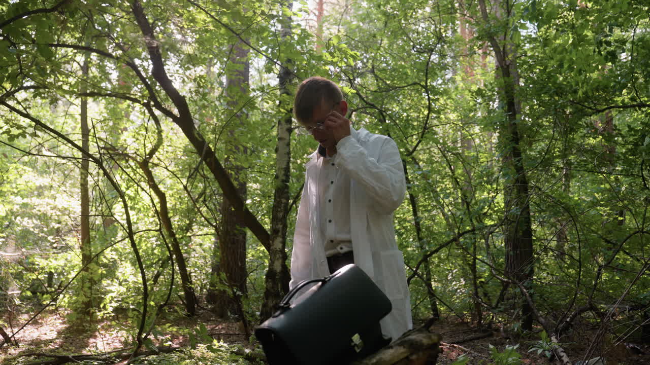 Young biologist wearing white lab coat carefully placing jotter into black backpack on old tree stump in forest under bright sunlight with green foliage and natural outdoor environment in background