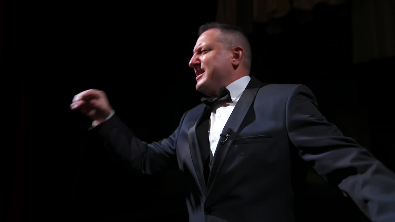 Actor in rich grey suit announces performance before auditorium. Theater curtains background. Dark background. Video from below.