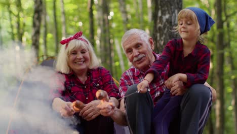 senior man woman with granddaughter resting at camping in wood cooking frying sausages over campfire