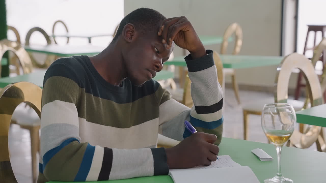 Black man writing after date note with wine glass on table, restaurant bistro setting with empty chairs, melancholic expression as he drafts apology and rereads memories, late evening mood of regret