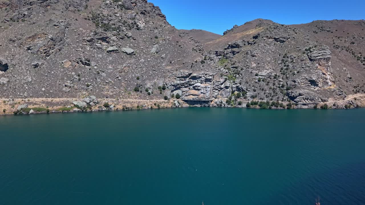 Drone flies forward on a sunny day, passing green trees, revealing the Clutha River, and approaching the Dunstan Trail winding through rocky hills and mountains