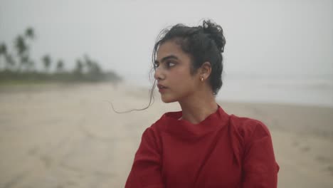 portrait of young indian female wearing red dress with nose ring on septum, looks to side turns head looking out side of camera with a beach in the background