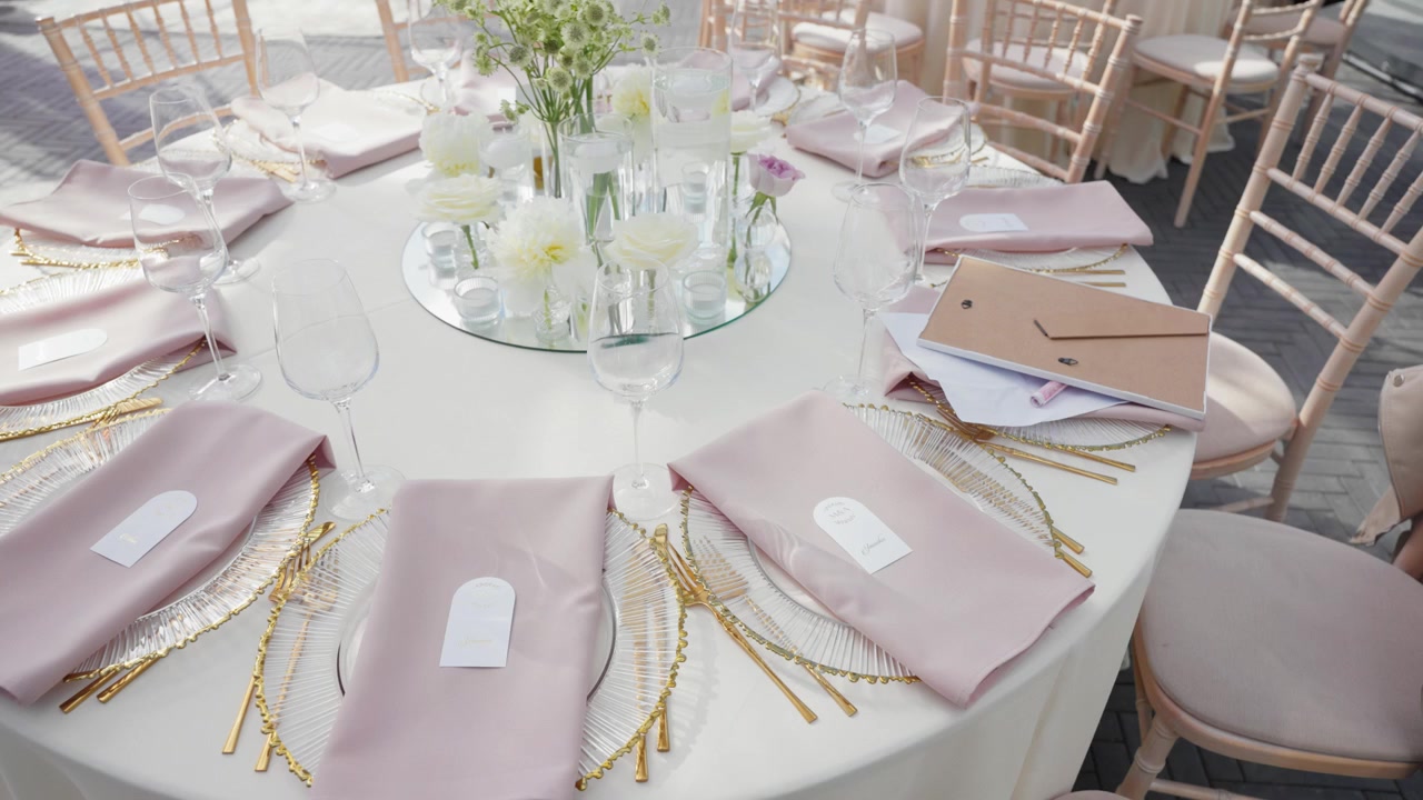 Beautiful pastel pink table set for an outdoor wedding reception with elegant napkins, gold accents, glassware, and fresh floral centerpiece. Soft natural light, inviting and romantic.