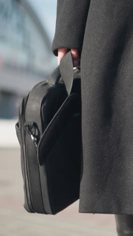 primer plano de una bolsa negra sostenida por una persona que lleva un elegante abrigo negro y botas de cuero, caminando por una calle urbana soleada con detalles borrosos de fondo de la arquitectura moderna y los peatones en movimiento