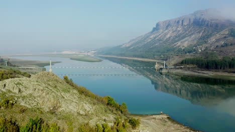 Aerial-view-of-a-river,-with-mountains-reflecting-in-the-water,-bridge-visible-on-a-sunny-day