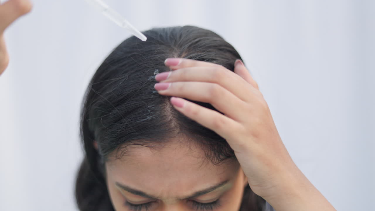 A young woman with a dropper applying hair serum on her hair root to improve hair growth in white background.