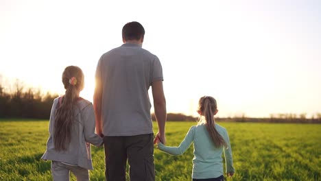 young, muscular man, father is holding his small daughters with hands. walking by the wide meadow. casual clothes. sun rays. footage from the backside