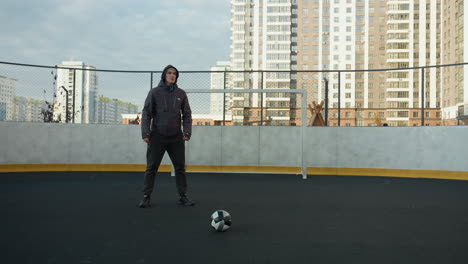 athlete performing squats with hands clasped in preparation for exercise on outdoor sports ground with soccer ball in foreground, surrounded by urban high-rise buildings and goal post