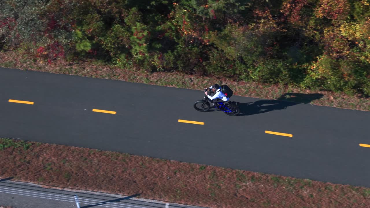 An aerial view of a man riding a bicycle on a paved bicycle path on a sunny day in autumn. The drone camera tilted downward, truck left to follow the man from his left side