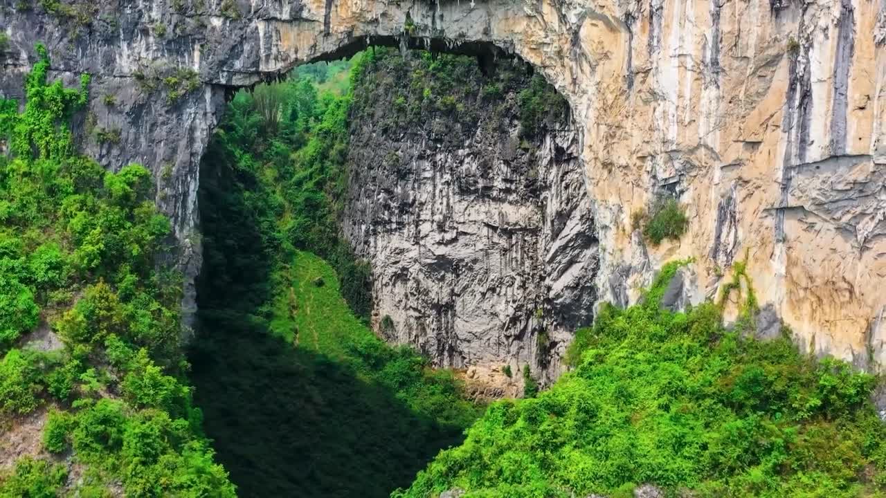 Aerial drone footage of the world deepest sinkhole in China that's also called Xiaozhai Tiankeng during a sunny day. Footage features a large hole in a cliff wall with green nature surrounding it.