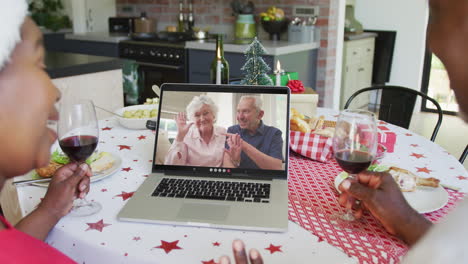 una pareja afroamericana con vino usando una computadora portátil para una videollamada de navidad con una pareja feliz en la pantalla