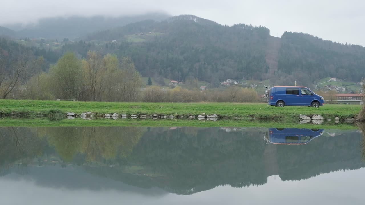 Blue volkwagon campervan driving on a overcast day. The camera panning from left to right with a beautiful reflection of a calm still lake. A couple travelling in a campervan