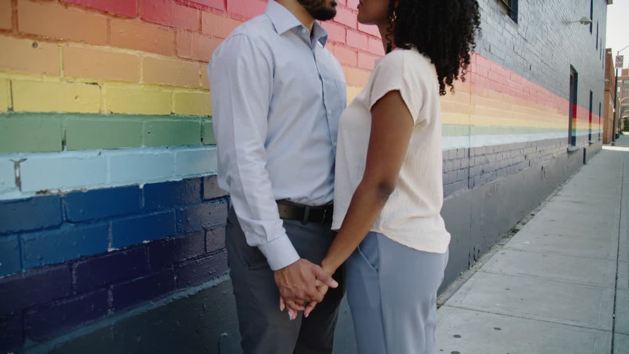 Couple en tenue de bureau, s'inclinant l'un vers l'autre, se tenant la main à la taille et se rapprochant sur un trottoir urbain orné d'une fresque arc-en-ciel.