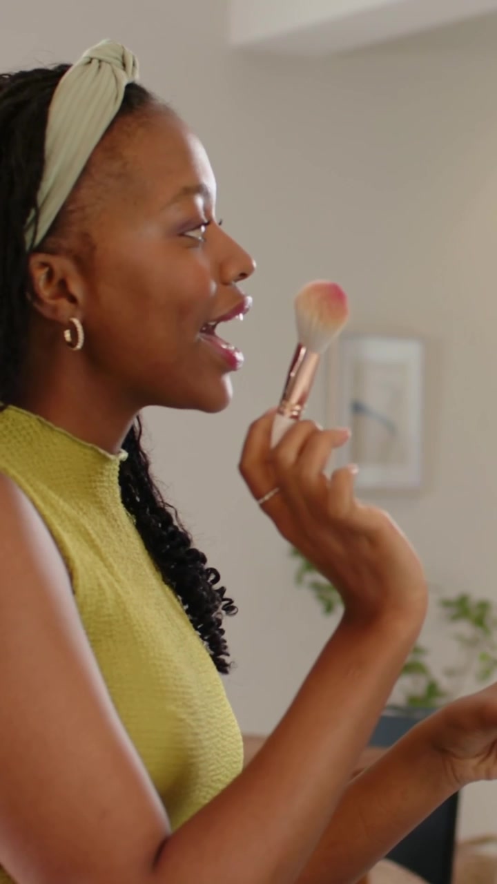 Vertical video: Applying woman in green top checking makeup at home vanity, holding fluffy brush