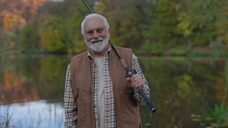 a smiling elderly man holding a fishing rod by a lake in autumn