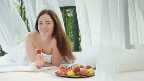 Mujer-Joven-En-La-Cama-Con-Una-Fresa-En-La-Mano-Yace-Junto-A-La-Piscina-En-Una-Tumbona-Con-Cortinas-Romance