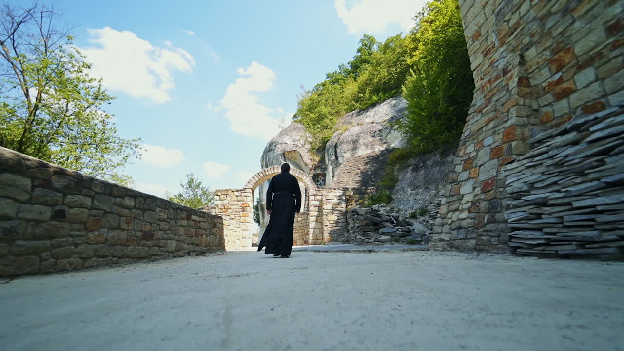 Monk wearing long black robe goes by the territory of rock monastery. Following the priest of orthodox church. Low angle view.