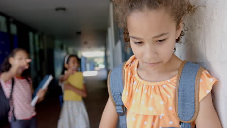 In school, girl with backpack feeling sad while other students pointing and laughing