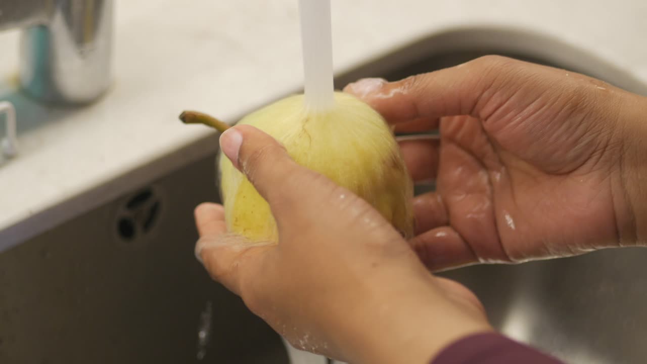 Manos lavando una pera fresca bajo el agua corriente en el fregadero de la cocina