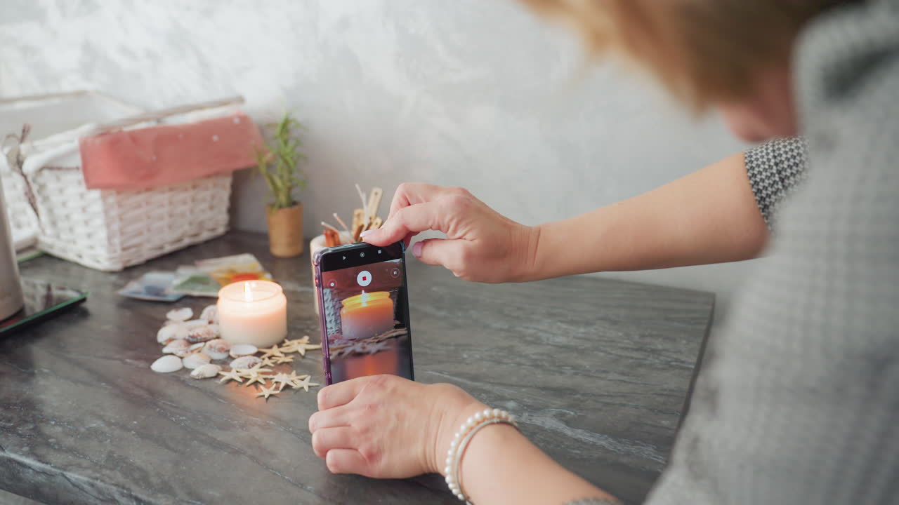 Videographer using smartphone to film candle arrangement on tabletop, demonstrating photography skill with steady hand movement while gliding phone above glowing candle surrounded by shells