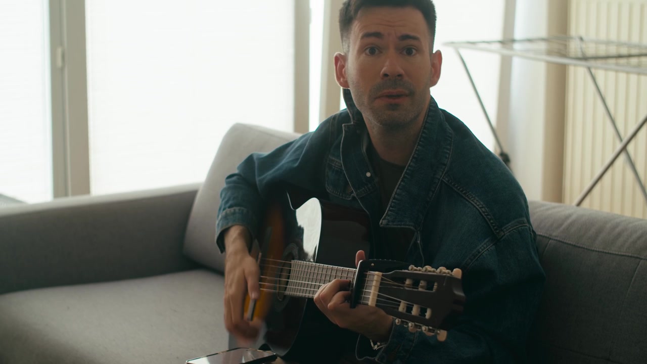 Una scena autentica di un uomo di talento che suona una chitarra classica mentre è seduto su un divano in un appartamento luminoso. Ottimo per contenuti di benessere e lifestyle