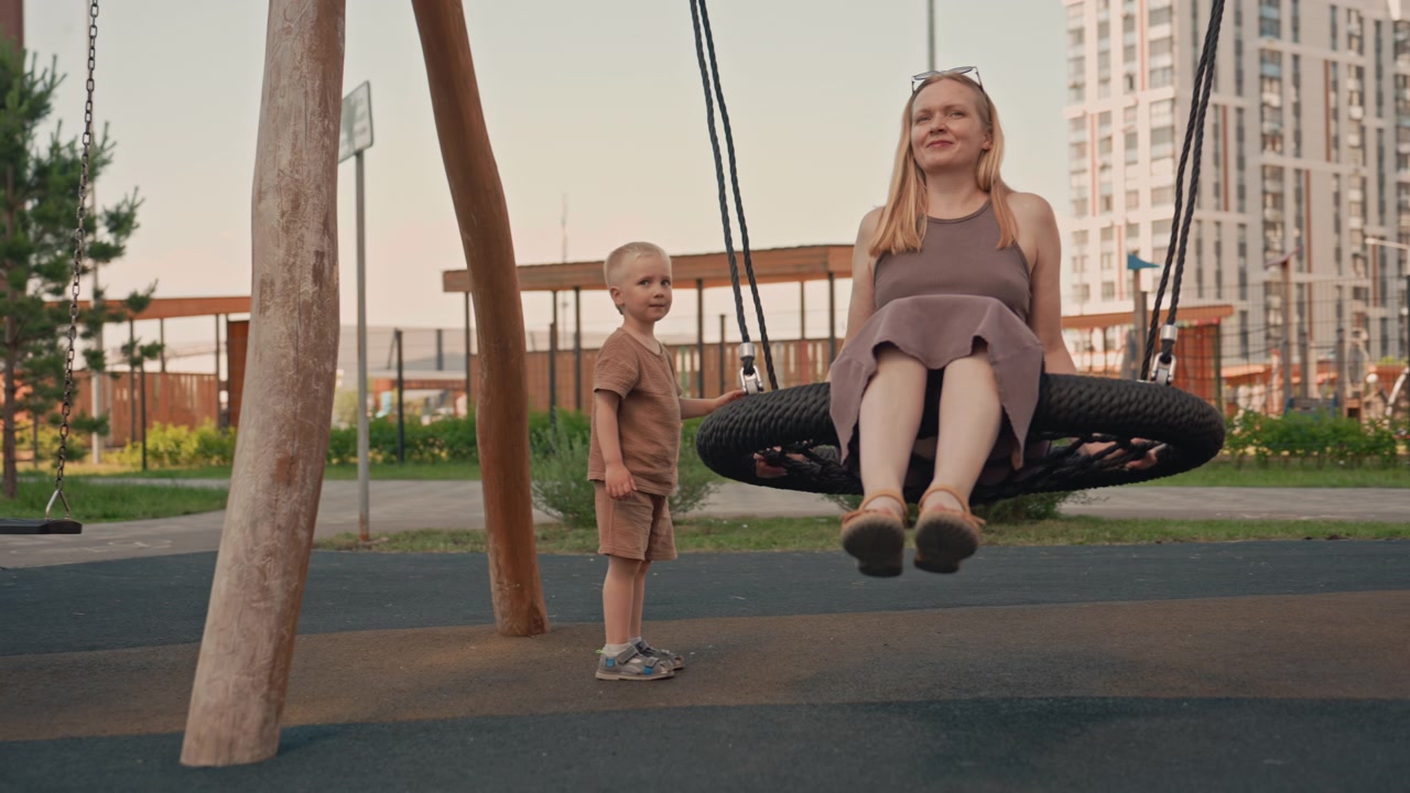 Donna e bambino interagiscono al parco giochi, con la donna seduta su un'altalena a pneumatico mentre il bambino sta in piedi lì vicino. La scena presenta attrezzature da gioco all'aperto su uno sfondo urbano di edifici sotto una luce soffusa.