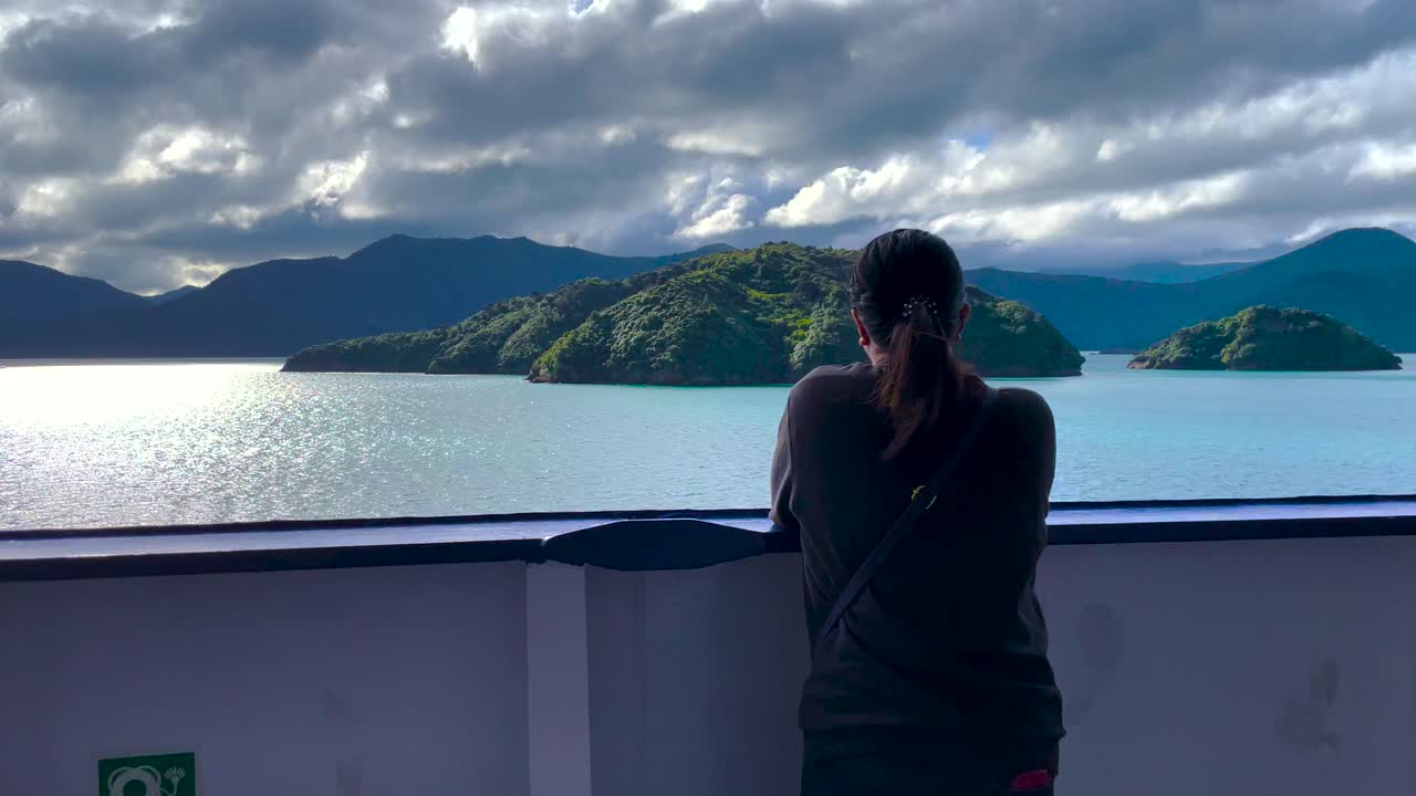 mujer de pelo oscuro en sudadera se apoya en la barandilla del barco y mira hacia las islas y el cielo nublado en la reina charlotte sound en nueva zelanda