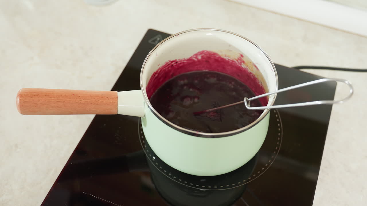 Close up of steaming berry sauce or compote bubbling inside mint green pot on black electric stove with wooden handle and metal whisk, showing intense heat and thick consistency during cooking