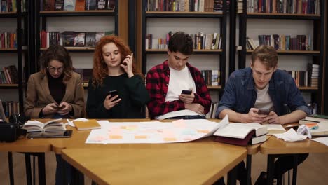 Group-of-four-university-students-two-males,-two-females-sitting-at-the-table-together-during-break-and-using-smartphones---texting,-surfing-internet-while-sitting-in-the-uni-library-back-to-the-bookshelves