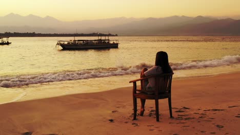 Indonesia-Island---A-Girl-Sitting-In-The-Wooden-Chair-Watching-The-Beauty-Of-Nature-Background-Anchored-Boats-In-The-Shallow-Ocean,-Lush-Island,-High-Mountains,-And-Foggy-Sky---Wide-Shot