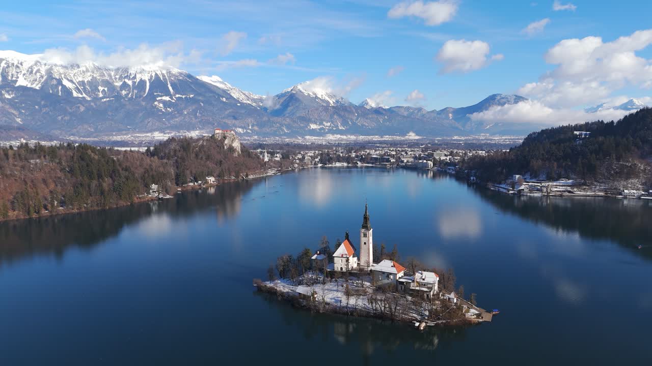 Vista aerea del lago di Bled in inverno, Slovenia, con l'iconica chiesa sull'isola circondata da acque blu calme, sponde innevate e montagne alpine sotto un cielo sereno e tranquillo. Campo largo