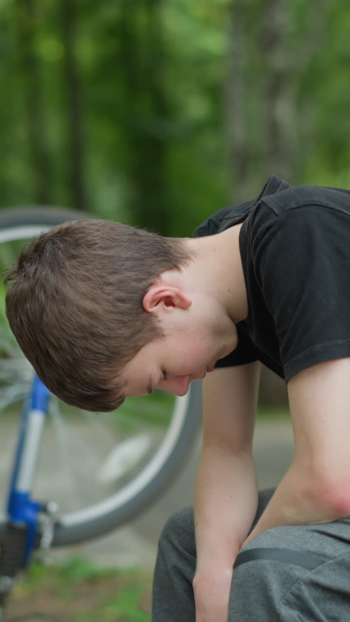 White boy in black top sits thoughtfully on park bench with his head down, his bicycle is upside down beside him, its tires still rotating, and the background features a blurred view of trees