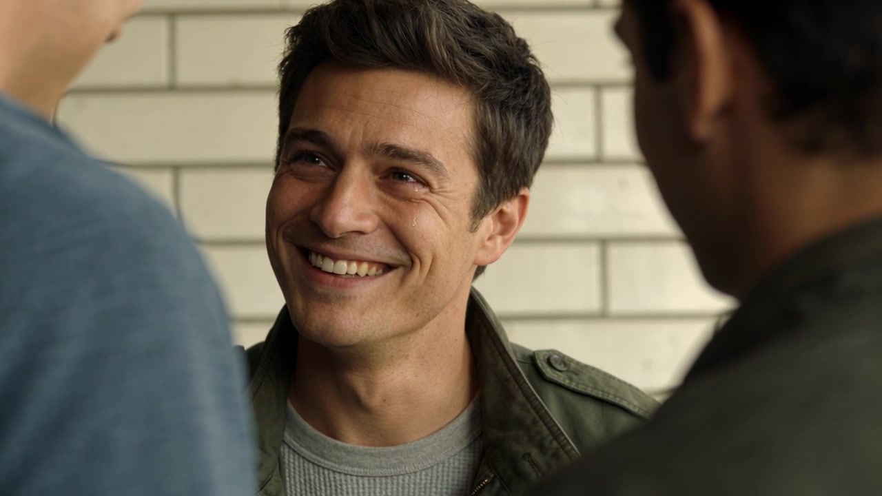Uomo sorridente annuisce, rispondendo all'uomo che parla alla sua sinistra, accanto a una parete di piastrelle chiare e con indosso una giacca verde oliva.