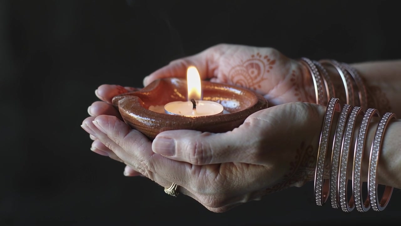 Entering frame, hennaed hands with bangles and ring holding clay diya at home, shielding flame