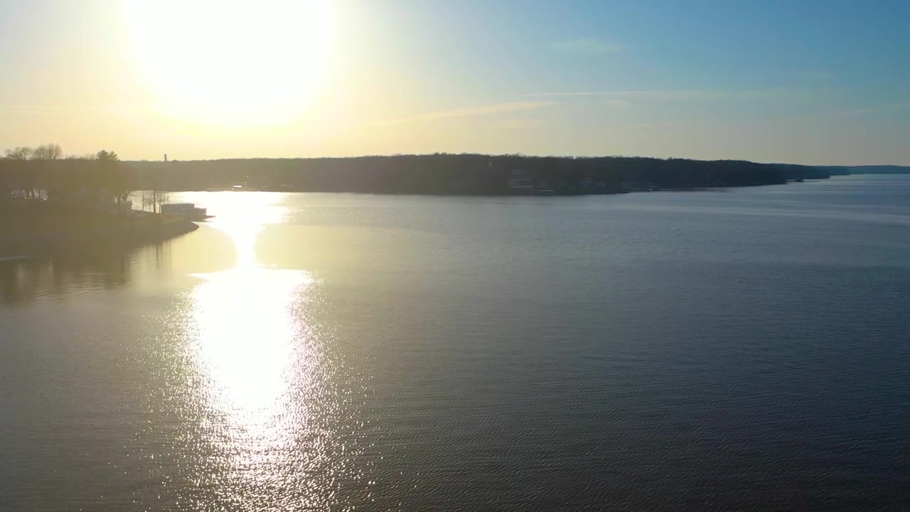 High drone view above Norris Lake, Tennessee at sunset, with shimmering reservoir water, wooded shoreline, distant houses, soft haze, and warm golden light across the calm surface.