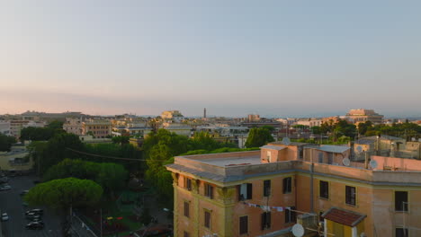 Low-flight-above-flat-roofs-with-TV-antennas-in-residential-urban-borough-at-golden-hour.-Revealing-lush-green-trees-in-park.-Rome,-Italy