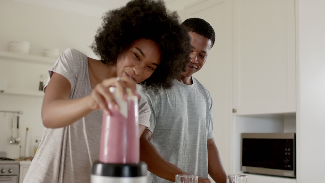 Menekan penutup, wanita mengoperasikan blender sementara pria menata gelas, menyiapkan smoothie merah muda untuk disajikan.